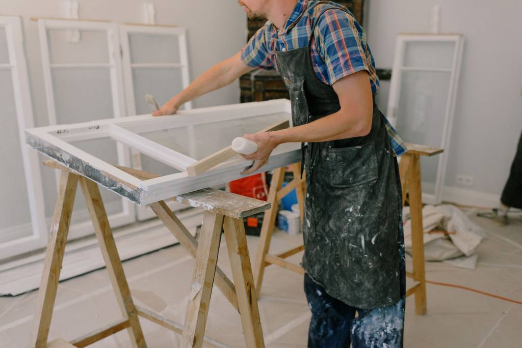 Decorator in a paint-spattered apron restoring an old timber window sash on trestles